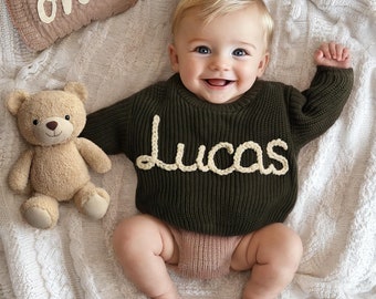 May include: A baby wearing a green knitted sweater with the name "Lucas" embroidered in white yarn. The baby is sitting on a white blanket with a brown pillow that says "one" on it.