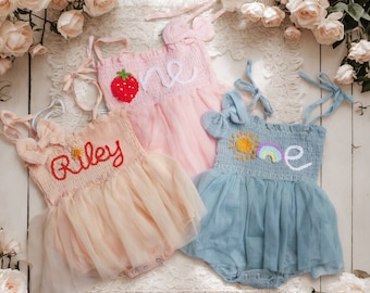 May include: Three tulle baby girl dresses with embroidered details. One dress is pink with a strawberry and the word "one" embroidered on it. Another dress is peach with the name "Riley" embroidered on it. The third dress is blue with a sun and rainbow and the word "one" embroidered on it.