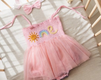May include: Pink baby dress with a tulle skirt and embroidered sun, rainbow, and the word "one". Includes a matching pink bow headband. The dress is on a neutral-colored crib mattress.
