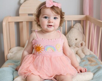 May include: A baby girl wearing a pink tulle dress with a rainbow and sun embroidery. She is sitting in a crib with a white stuffed animal beside her. She is wearing a pink headband with a bow.