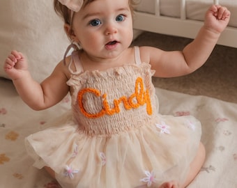 May include: A baby girl wearing a light pink tulle dress with white flowers embroidered on it. The dress has a cream-colored bodice with the name "Cindy" embroidered in orange yarn. The baby is wearing a light pink velvet bow in her hair.