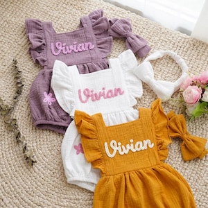 Könnte beinhalten: Drei Baby-Strampler in Lila, Weiß und Senfgelb, jeweils mit dem Namen "Vivian" bestickt. Die Strampler haben Rüschendetails und werden von passenden Accessoires wie einem Stirnband und Schleifen begleitet.