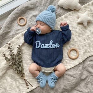 Puede incluir: Un bebé con un suéter de punto azul marino con el nombre "Daxton" en escritura azul claro. El bebé también lleva un gorro de punto azul claro con un pompón, botines a juego y un chupete. El bebé está acostado sobre una manta beige.