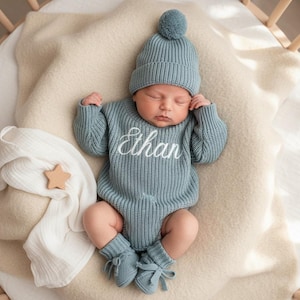 May include: A newborn baby wearing a light blue knitted outfit, including a hat with a pom-pom, a long-sleeved romper with the name "Ethan" embroidered in white, and matching booties. The baby is lying on a cream-colored blanket.