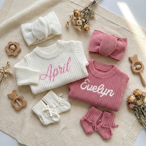 May include: A collection of baby clothing and accessories in a soft, neutral palette. Includes a white sweater with "April" embroidered in pink, a pink sweater with "Evelyn" embroidered in white, matching headbands, and booties. Wooden toys and dried flowers complete the arrangement.