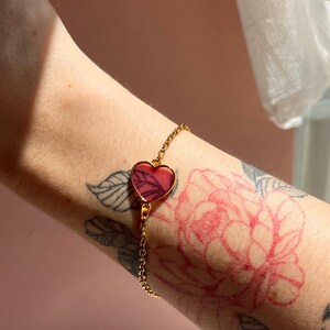 Peut inclure: Un bracelet en chaîne dorée avec un pendentif en forme de cœur rose. Le pendentif en forme de cœur est translucide et présente un design délicat.