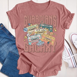 Op de afbeelding: Een roze T-shirt met een grafisch ontwerp van cassettebandjes en de tekst "Guardians of the Galaxy". De cassettebandjes hebben verschillende songtitels, waaronder "September", "Conga", "One Way", "Disco Inferno", "Everybody Wants to Rule the World" en "I Ran".