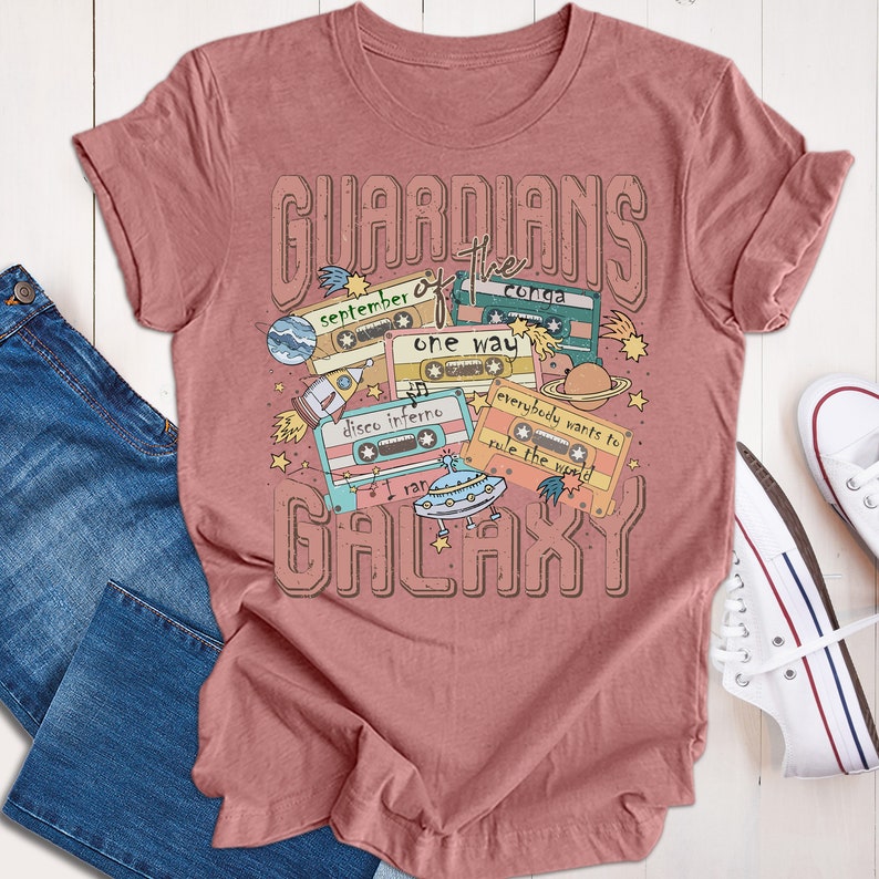 Op de afbeelding: Een roze T-shirt met een grafisch ontwerp van cassettebandjes en de tekst "Guardians of the Galaxy". De cassettebandjes hebben verschillende songtitels, waaronder "September", "Conga", "One Way", "Disco Inferno", "Everybody Wants to Rule the World" en "I Ran".