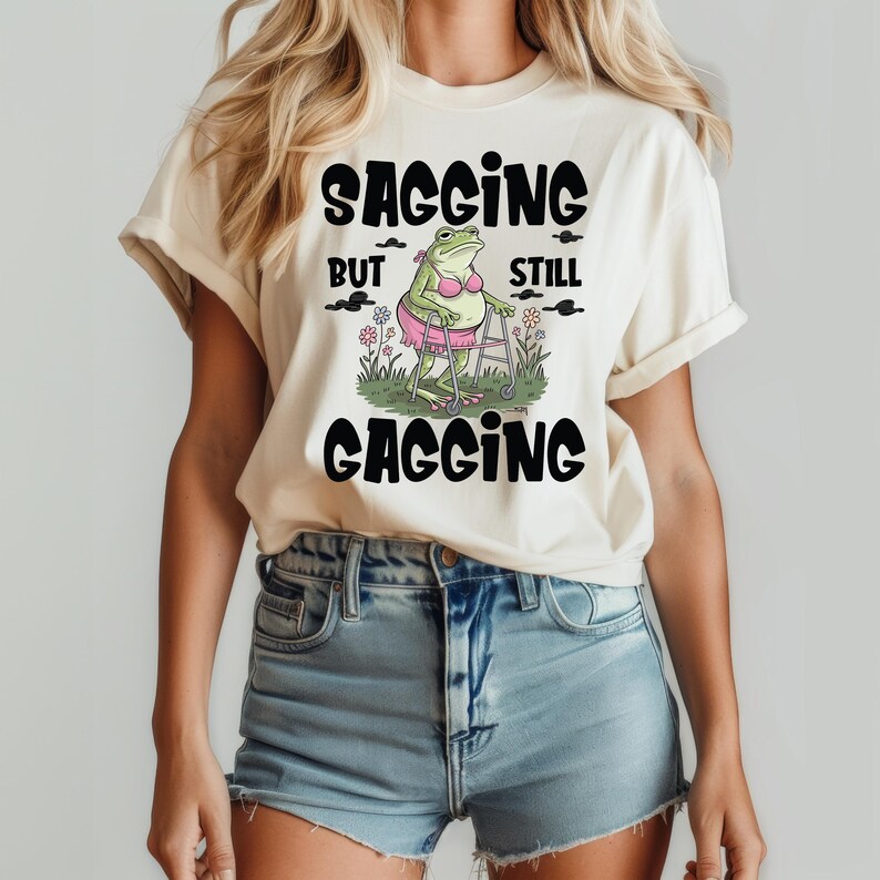 May include: A white t-shirt with a black graphic of a frog wearing a pink bra and using a walker. The text on the shirt reads "Sagging But Still Gagging".