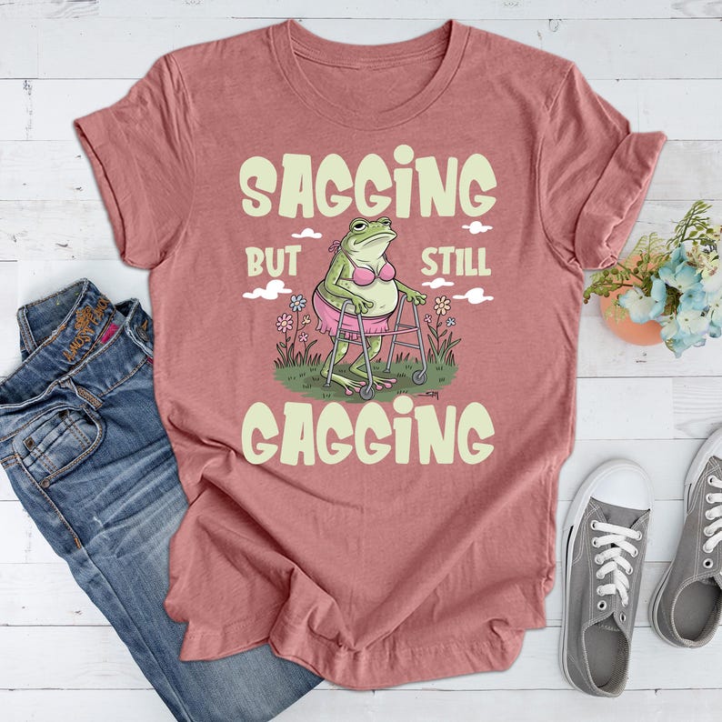 May include: A pink t-shirt featuring a green frog wearing a pink bikini and using a walking frame. The text on the shirt reads "Sagging but still gagging".
