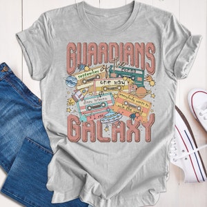 Op de afbeelding: Een grijs T-shirt met een grafisch ontwerp van cassettebandjes en sterren. De tekst "GUARDIANS of the GALAXY" is in roze op het shirt gedrukt. De cassettebandjes hebben titels zoals "September", "One Way", "Disco Inferno" en "Everybody Wants to Rule the World".