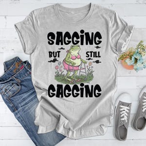 May include: Grey t-shirt featuring a cartoon frog in a pink bikini using a walking frame. The text on the shirt reads "Sagging but still gagging".