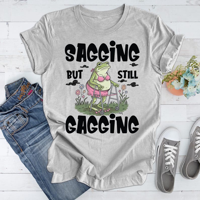 May include: Grey t-shirt featuring a cartoon frog in a pink bikini using a walking frame. The text on the shirt reads "Sagging but still gagging".