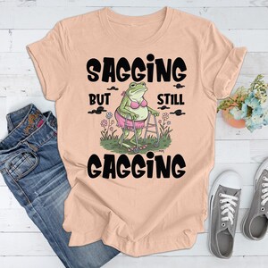 May include: A light peach t-shirt featuring a graphic of a frog wearing a pink bikini and using a walker. The text "Sagging But Still Gagging" is printed on the shirt.