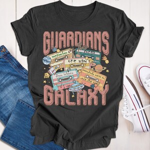 Op de afbeelding: Een donkergrijs T-shirt met een retro ontwerp met de tekst "Guardians of the Galaxy" met cassettebandjes en een UFO. De cassettebandjes hebben songtitels zoals "September", "Conga", "One Way", "Disco Inferno" en "Everybody Wants to Rule the World".
