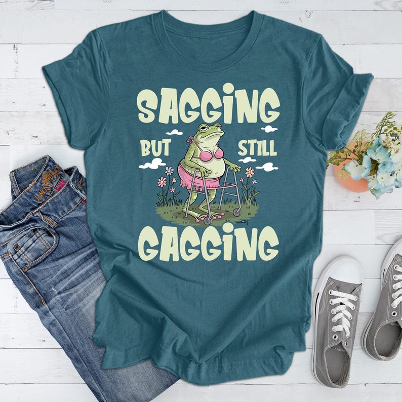 May include: Teal blue t-shirt featuring a green frog wearing a pink bikini and using a walking frame. The text on the shirt reads "Sagging But Still Gagging".