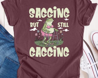 May include: A maroon t-shirt featuring a green frog wearing a pink bra and using a walking frame. The text on the shirt reads "Sagging but still Gagging".