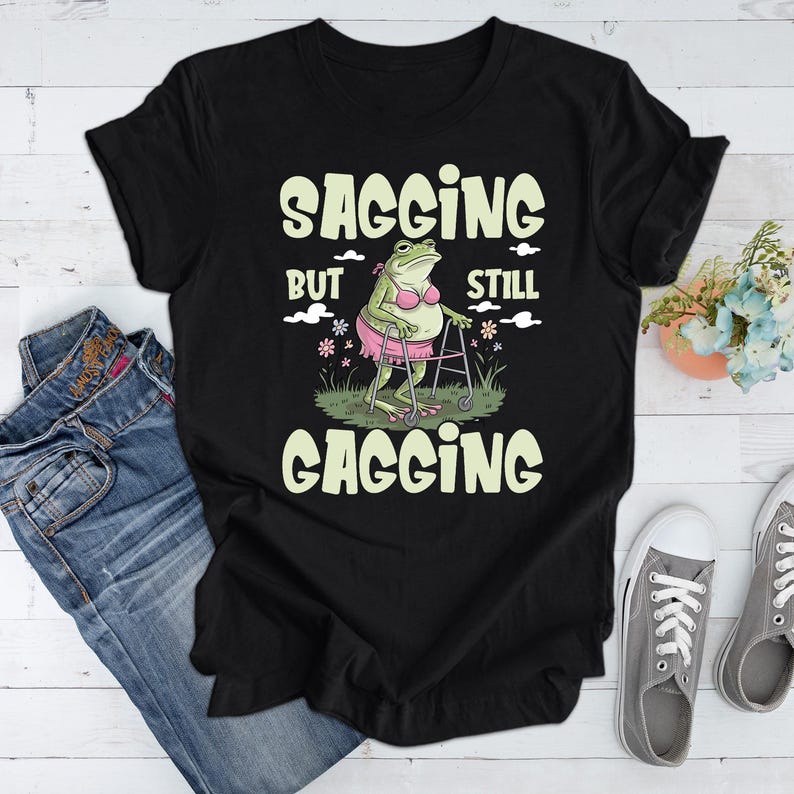 May include: Black t-shirt featuring a green frog wearing a pink bikini and using a walking frame. The text on the shirt reads "Sagging but still gagging."
