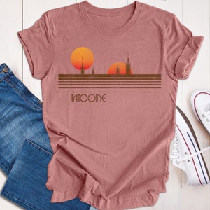 May include: Dusty rose t-shirt featuring a Tatooine graphic. The design includes two orange suns, a brown skyline, and the word "TATOOINE" in a stylized font. The shirt is paired with blue jeans and white sneakers.