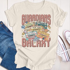 Op de afbeelding: Een wit T-shirt met een roze grafisch ontwerp met de tekst "Guardians of the Galaxy" met een retro cassettebandthema. De grafiek bevat cassettebandjes met songtitels zoals "September", "Conga", "One Way", "Disco Inferno", "Everybody Wants to Rule the World" en "I Ran".