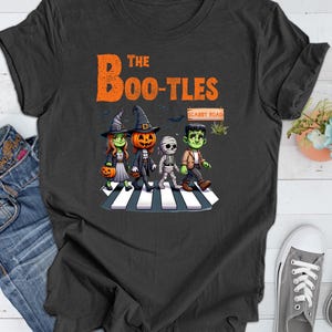 May include: A dark gray t-shirt with a graphic of four Halloween characters walking across a crosswalk. The characters are a witch, a pumpkin, a mummy, and a Frankenstein. The text "The Boo-Tles" is above the characters and "Scabby Road" is above the crosswalk.