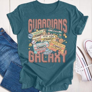Op de afbeelding: Tealblauwe T-shirt met een grafische print van cassettebandjes en de tekst "Guardians of the Galaxy". De cassettebandjes hebben songtitels zoals "September", "Conga", "One Way", "Disco Inferno", "I Ran" en "Everybody Wants to Rule the World".