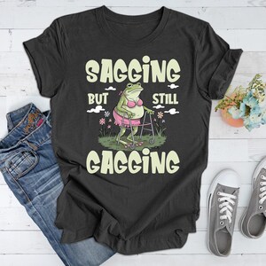 May include: A dark grey t-shirt featuring a green frog wearing a pink bikini and using a walking frame. The text on the shirt reads "Sagging but still gagging".