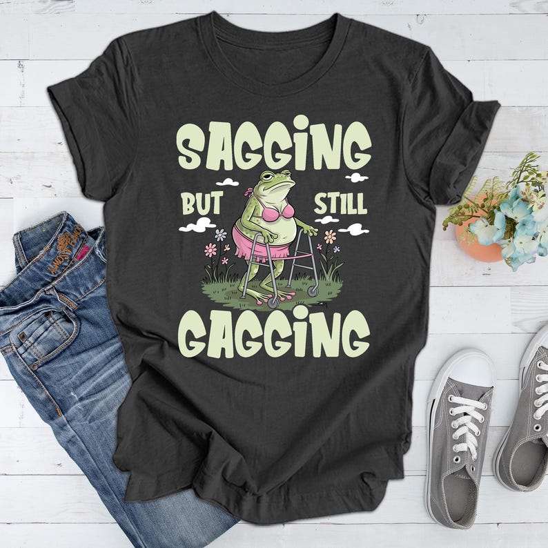 May include: A dark grey t-shirt featuring a green frog wearing a pink bikini and using a walking frame. The text on the shirt reads "Sagging but still gagging".