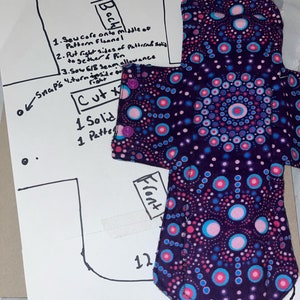 May include: A purple fabric menstrual pad with a pink and blue polka dot pattern. The pad is folded in half and has a white label that says "Front". The pad is on a white piece of paper with a hand-drawn diagram of the pad and instructions for sewing it.
