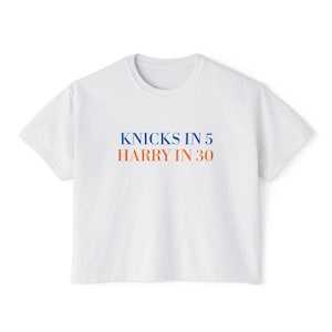 May include: A white short-sleeved t-shirt featuring the text KNICKS IN 5 in blue letters on the top line and HARRY IN 30 in orange letters on the bottom line, centered on the chest of the garment.