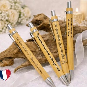 Peut inclure: Quatre stylos en bambou avec des clips et des pointes en métal argenté. Chaque stylo présente des textes et des graphiques gravés au laser, incluant des dates, des noms et des phrases comme 'merci de m'avoir aidé a grandir' et 'A2TP ENTREPRISE DU BÂTIMENT À BLAN'.