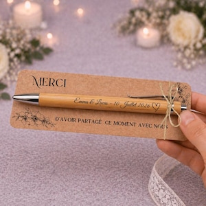 Pode incluir: Uma caneta esferográfica de bambu com detalhes prateados, gravada com o texto Emma & Lucas - 10 Juillet 2026 e um símbolo de coração, presa a um cartão de papel kraft retangular impresso com MERCI e D'AVOIR PARTAGÉ CE MOMENT AVEC NOUS.