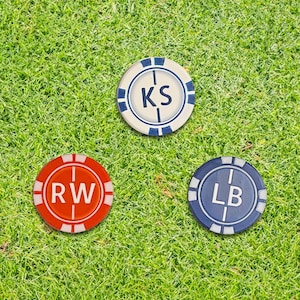 May include: A white golf ball and three personalized golf ball markers on green grass. The markers are red, blue, and off-white, each with initials. The grass is a vibrant green.