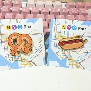 May include: Two enamel pins on cards. One pin is a pretzel with a rat design, the other is a hot dog with a rat design. The cards have a subway map design and the text "NYC Rats".