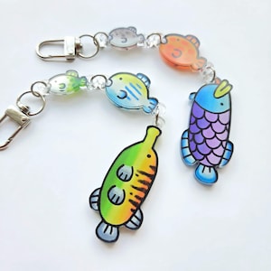 May include: Two silver-toned metal lobster clasps attached to chains featuring multiple colorful acrylic fish charms. The charms include a green and orange fish, a blue fish, a purple and blue fish, and a small grey fish, all with black outlines.