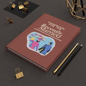 May include: A brown hardcover book with the title "Handbook for the Recently Married" in white text. The cover features a retro illustration of a couple walking away from the viewer, with a sun and clouds in the background.