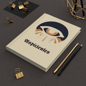 Puede incluir: Una libreta beige con un gráfico de ojo estilizado y la palabra "Uspicientes" en una fuente decorativa.