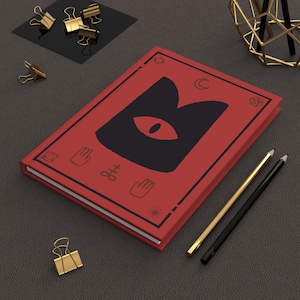 Pode incluir: Um diário de capa dura vermelho com uma ilustração de um gato preto e vários símbolos na capa. O gato tem um olho vermelho grande. O diário está sobre uma superfície cinza escura com dois lápis e um suporte decorativo de metal dourado.