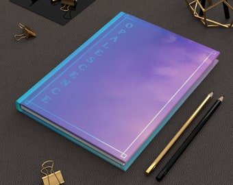 Op de afbeelding: Een paars en blauw notitieboek met een witte rand en de tekst "ECZECSEAPO" op de omslag. Het notitieboek ligt op een grijs, getextureerd oppervlak met twee potloden en een gouden paperclip.