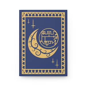 Peut inclure: Un carnet bleu marine avec une bordure dorée et un motif de croissant de lune. La lune a un motif floral stylisé et un cercle avec un motif géométrique à l'intérieur. Il y a quatre étoiles dorées autour de la lune.