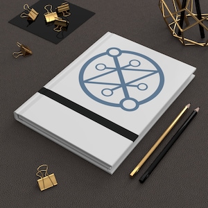 May include: White notebook with a black elastic band and a blue geometric symbol on the cover. Two pencils, one gold and one black, are lying on a dark brown surface next to the notebook.