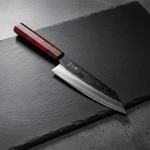 Puede incluir: Un cuchillo de cocina de estilo japonés con una hoja oscura y texturizada y un mango de madera con detalles rojos y marrones. El cuchillo está inclinado sobre una superficie oscura, mostrando su filo y artesanía.