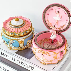 May include: Two decorative music boxes. One is pink and blue with a carousel design. The other is pink with a ballerina and swan inside. The lid is open, revealing a ballerina figurine. The boxes sit on top of books.