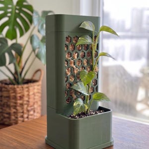 May include: A dark olive green vertical plant support structure featuring a rectangular base and a tall, hollow column with a honeycomb-patterned front panel filled with light brown sphagnum moss, supporting a green and yellow variegated pothos plant.