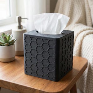 May include: A black tissue box with a honeycomb pattern sits on a wooden table. White tissues are visible at the top. A succulent in a white pot and a soap dispenser are in the background.