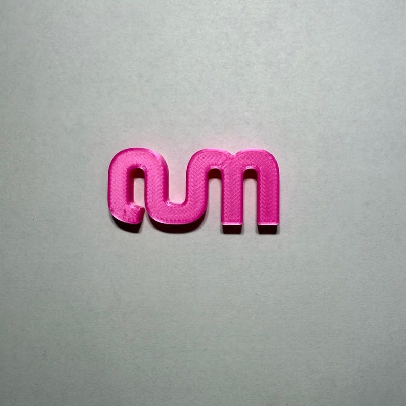 May include: A bright pink, three-dimensional plastic object featuring the lowercase letters 'nm' connected in a continuous, rounded, sans-serif font. The surface of the letters displays a visible, fine, horizontal 3D-printed texture.