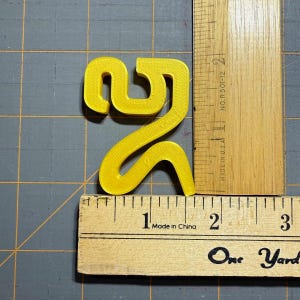 May include: A bright yellow, 3D-printed plastic numeral two, measuring approximately 6.4 cm in height and 5.1 cm in width, positioned next to wooden rulers that show the scale in centimetres on a grey cutting mat with a grid pattern.