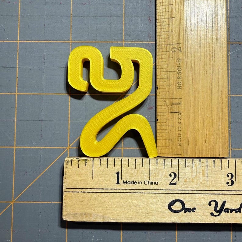 May include: A bright yellow, 3D-printed plastic numeral two, measuring approximately 6.4 cm in height and 5.1 cm in width, positioned next to wooden rulers that show the scale in centimetres on a grey cutting mat with a grid pattern.