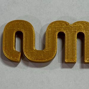 May include: A three-dimensional, gold-coloured plastic object featuring the lowercase letters 'nm' connected in a continuous, flowing script. The surface displays horizontal ridges characteristic of a 3D-printed manufacturing process.