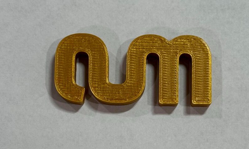May include: A three-dimensional, gold-coloured plastic object featuring the lowercase letters 'nm' connected in a continuous, flowing script. The surface displays horizontal ridges characteristic of a 3D-printed manufacturing process.